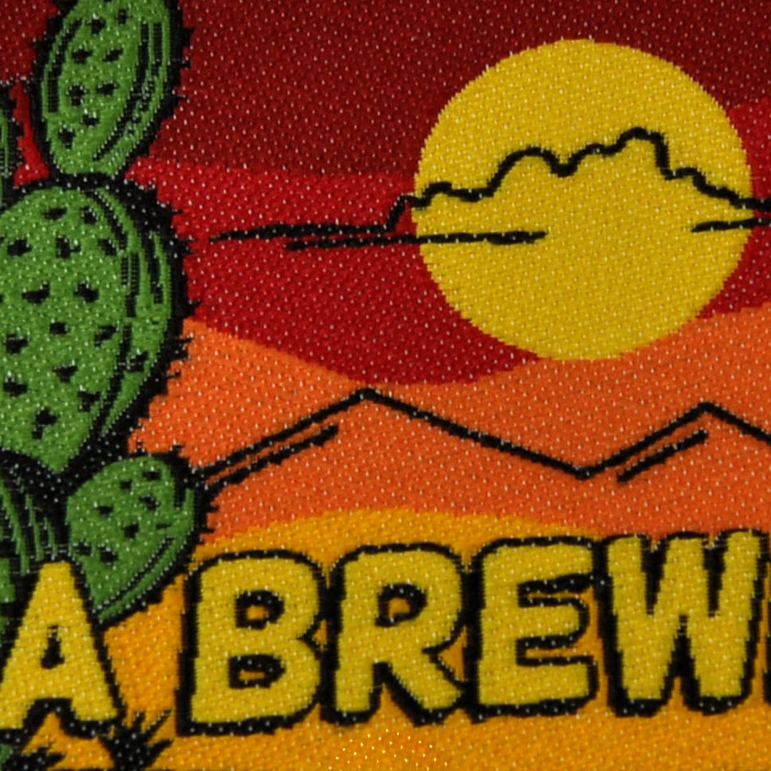 Close-up of a woven patch featuring a desert scene with a cactus, mountains, a setting sun, and partial text
