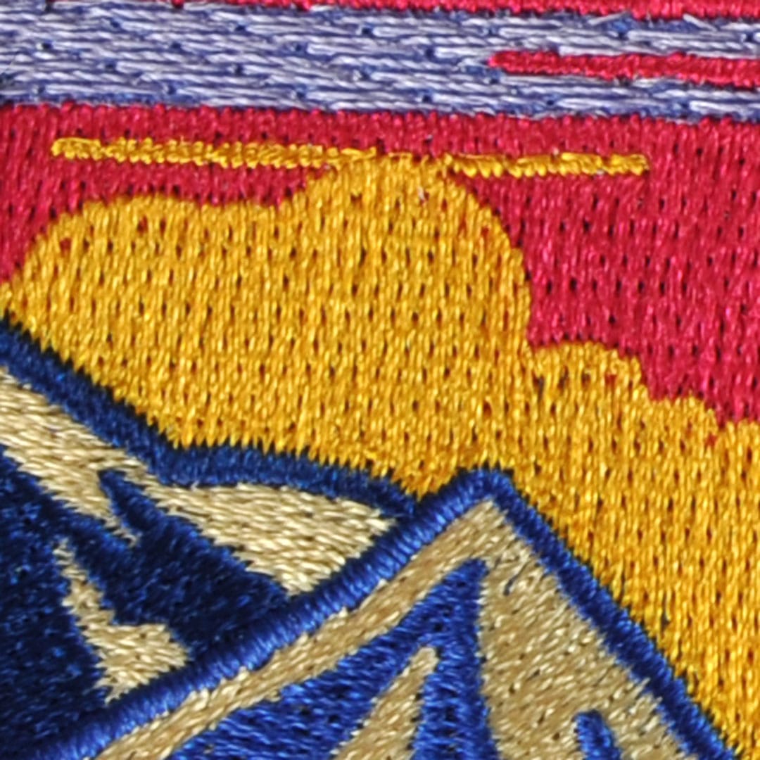 Close-up of an embroidered patch showing complex-fill and satin stitches, featuring a landscape with mountains, clouds, and a colorful sky
