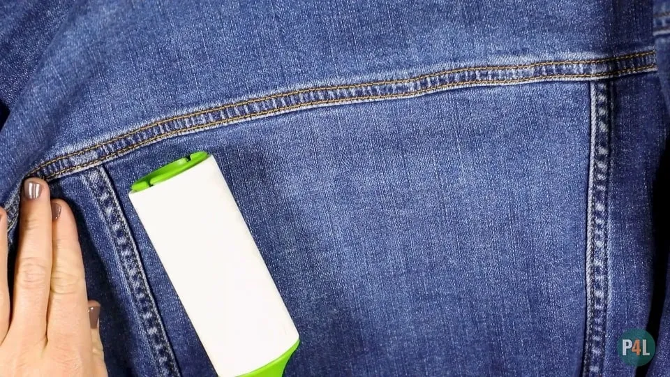 Hand holding a lint roller on the back of a denim jacket, preparing the fabric for iron-on patches.