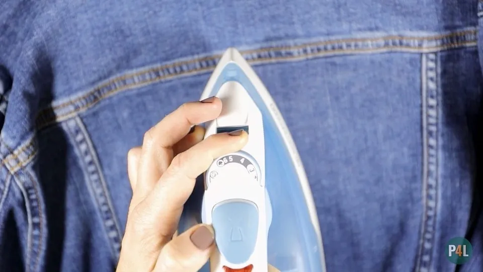 Hand adjusting the temperature settings on an iron, with a denim jacket in the background, preparing to apply iron-on patches.