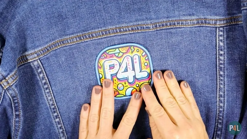 Hands positioning a colorful iron-on patch that reads 'P4L' onto the back of a denim jacket.