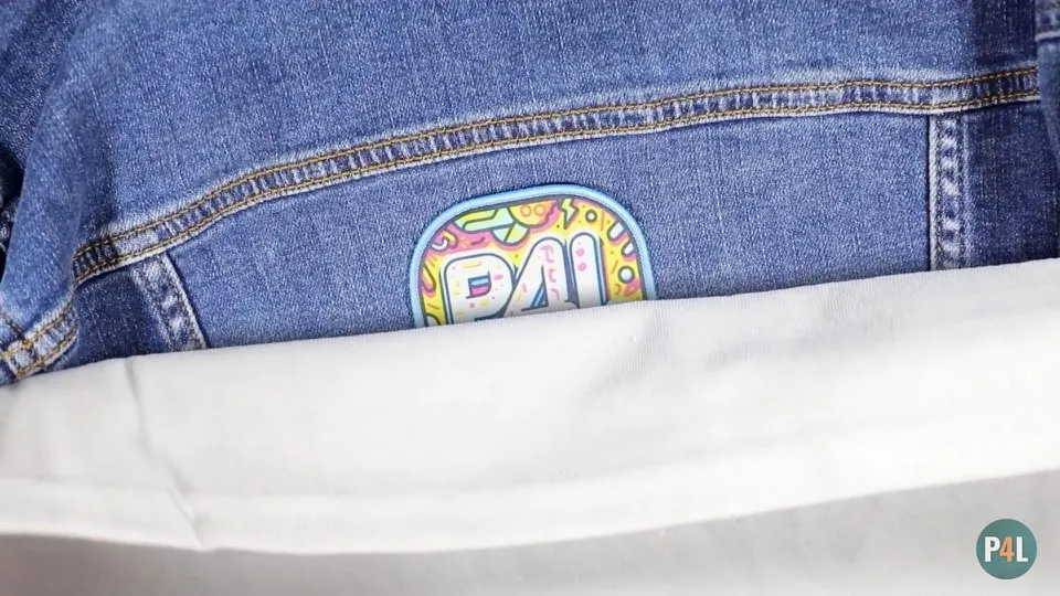 Denim jacket with a colorful 'P4L' iron-on patch partially covered by a white press cloth, preparing to iron the patch.