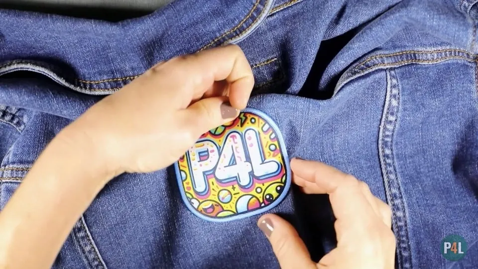 Hands checking the adhesion of a colorful 'P4L' iron-on patch on the back of a denim jacket.