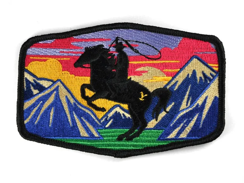 Embroidered patch featuring a silhouette of a cowboy on a rearing horse with a lasso, set against a colorful mountain and sunset background.