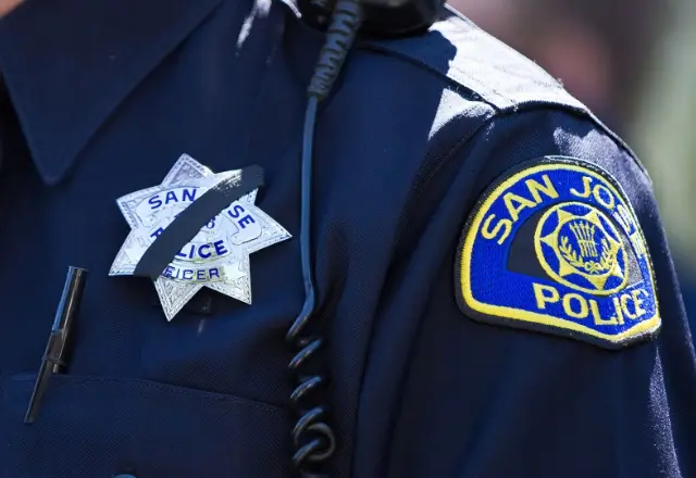 Close-up of a San Jose Police officer’s uniform, showing a silver badge with a black mourning band across it and a blue and yellow San Jose Police patch on the shoulder.